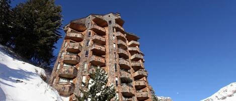 Un edificio alto de varios pisos con numerosos balcones, rodeado de montañas y árboles cubiertos de nieve.