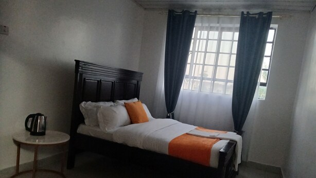 A single bed with a wooden headboard, a small round table with a kettle, and a window with curtains. - Rhacky Homes Syokimau | Near JKIA 5 - 10 minutes' drive | Free WI-FI & Parking. (Machakos)