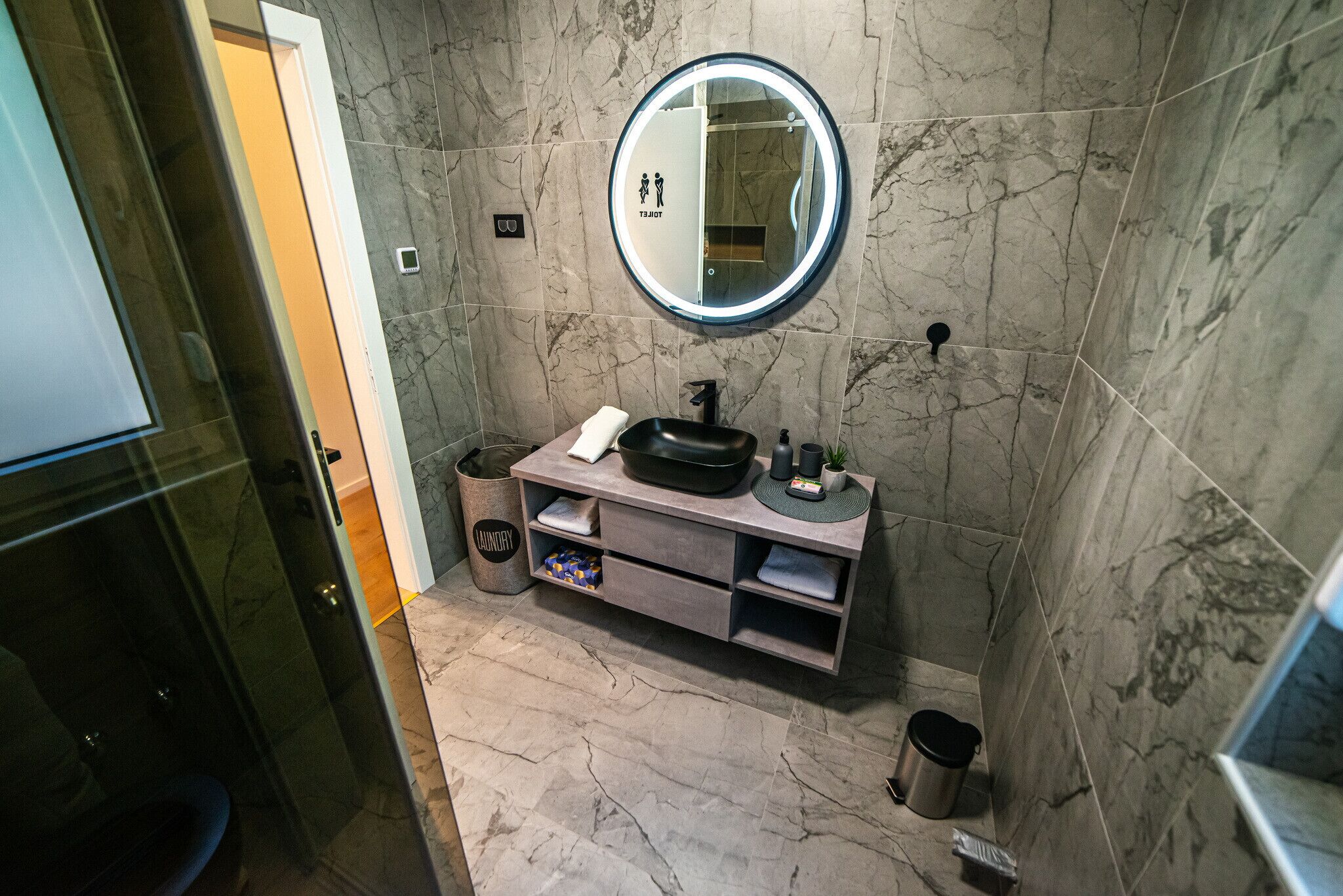 A modern bathroom with a marble floor, a round mirror with a light, a sink with a black basin, and a towel rack.