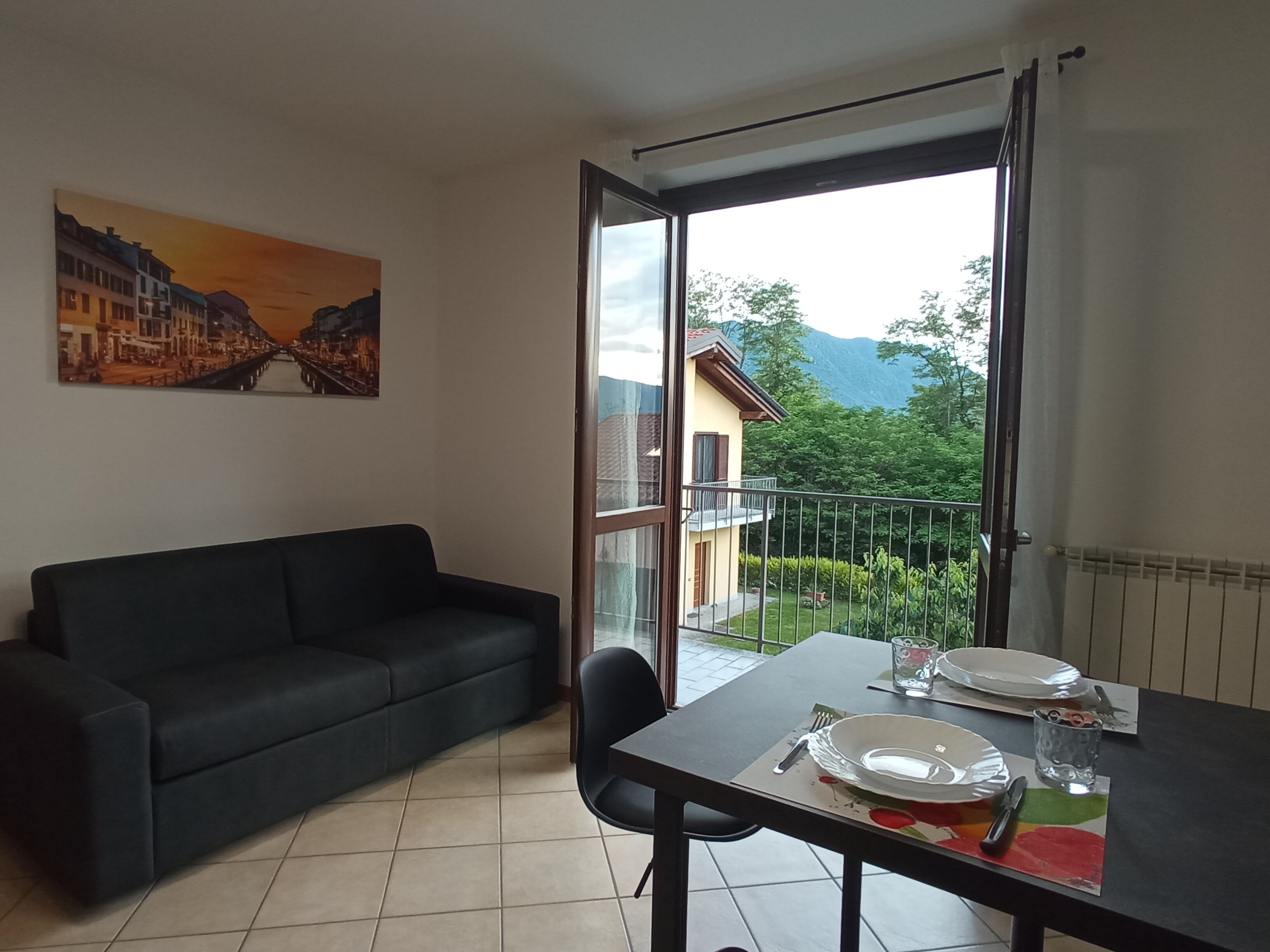 A living room with a black sofa, a dining table with plates and cutlery, and a large window offering a view of a mountain and greenery.