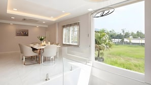 A modern dining area with a glass partition, white furniture, and a view of a green landscape. - Bangkok's Most Beautiful Modern Villa (Bangkok)