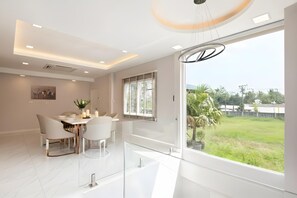 A modern dining area with a glass partition, white furniture, and a view of a green landscape. - Bangkok's Most Beautiful Modern  Villa (Bangkok)