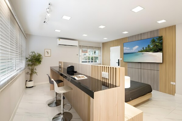 A modern hotel room with a kitchen area, a bed, and a large TV displaying a beach scene.