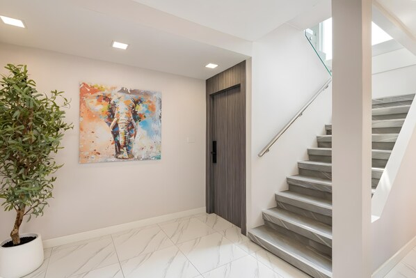 A modern hallway with a marble floor, a potted plant, a colorful abstract painting, a wooden door, and a staircase. - Bangkok's Most Beautiful Modern Villa (Bangkok)