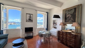 A living room with a TV, dining table, and chairs, large windows with a view of the sea, and a wooden cabinet with a lamp.
