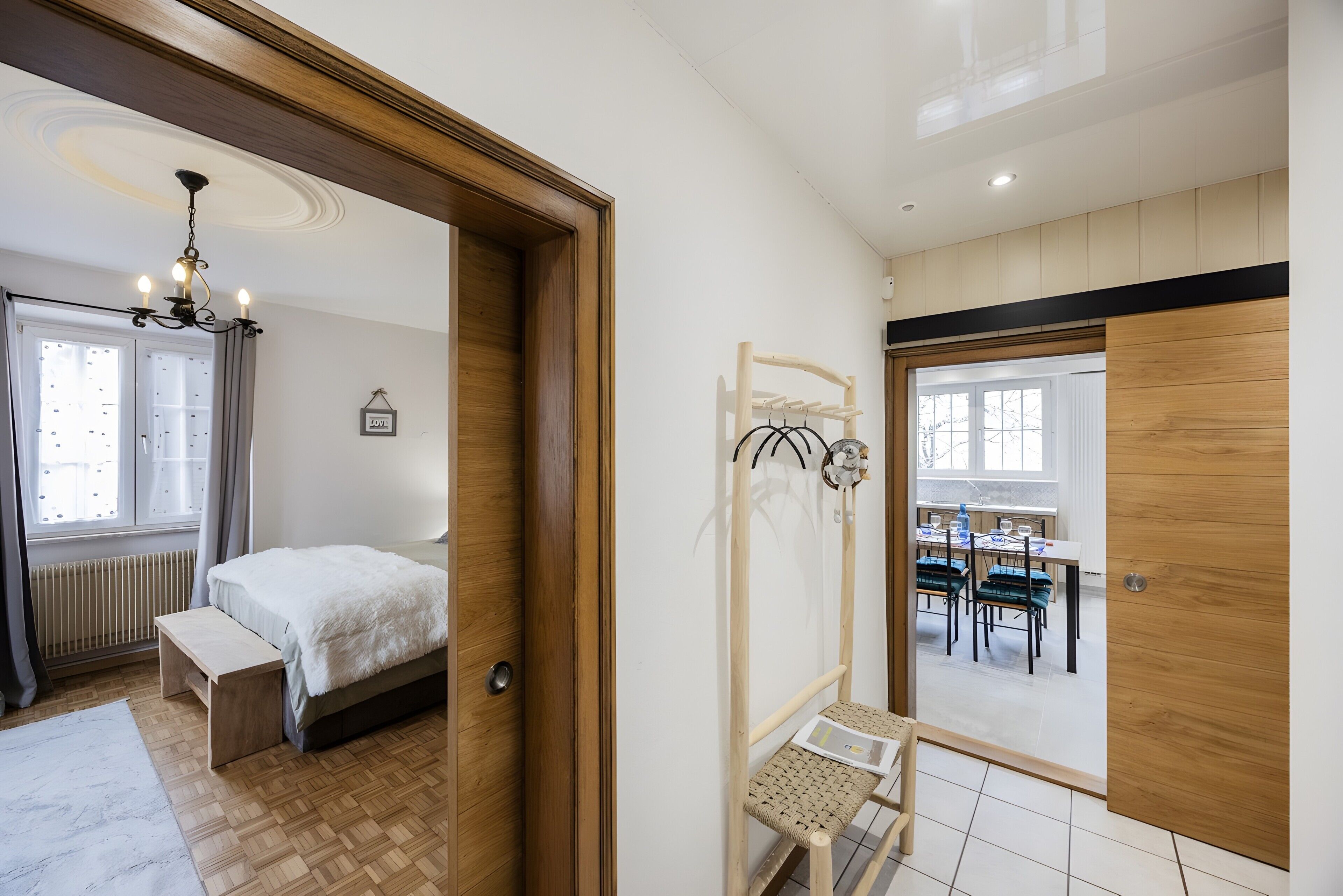 Een slaapkamer met een bed, nachtkastje, bankje, houten deur en uitzicht op een eetruimte.