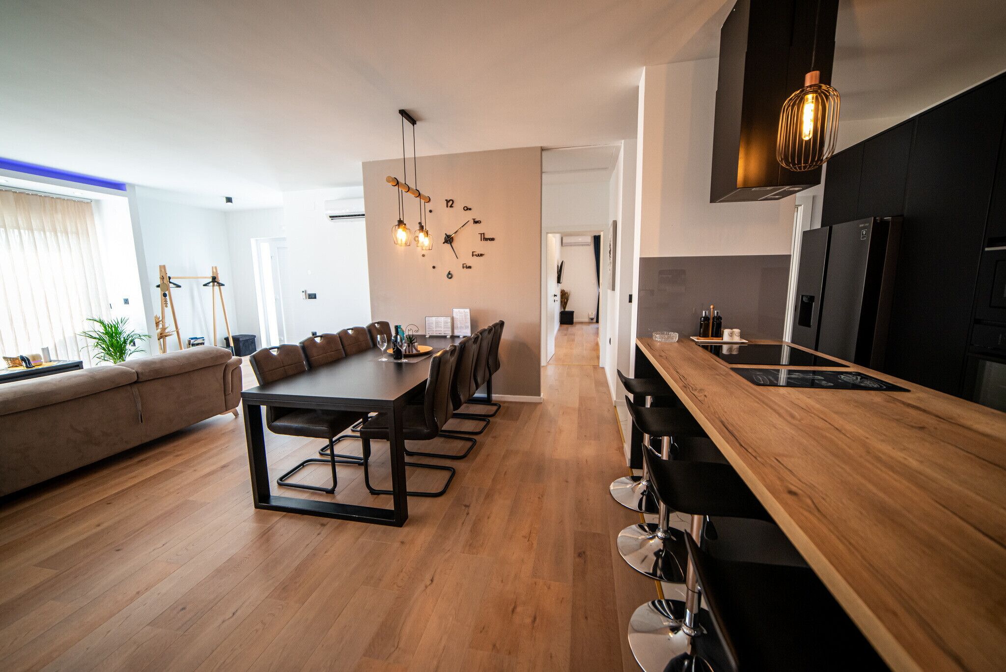 Una cucina moderna con piano di lavoro in legno e mobili neri, una zona pranzo con un tavolo lungo e sedie, e un soggiorno con divano e pianta.