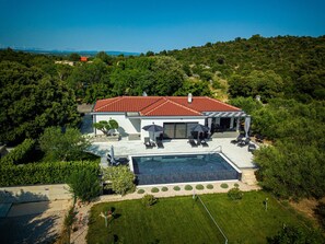 Ein modernes Haus mit Schwimmbad, umgeben von üppiger Vegetation und Blick auf Windkraftanlagen in der Ferne.