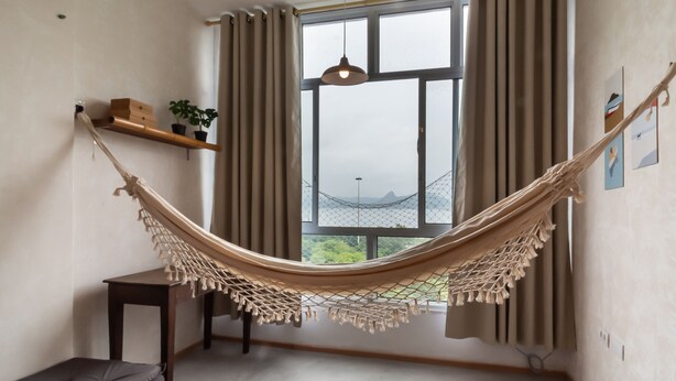 A room with a hammock, a small table, a bench, a shelf with plants, and a large window with curtains. - Ocean and Sugarloaf Mountain View Prf805 (Rio de Janeiro)
