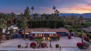 A modern house with a flat roof, surrounded by palm trees and red flowering bushes. - Remodeled Palm Desert Home by El Paseo: Mountain views, pool&spa, outdoor kitchen, sleeps 8 (Palm Desert)