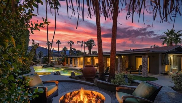 A luxurious outdoor setting with a fire pit, lounge chairs, a swimming pool, and a well-lit building with stone and glass architecture. - Remodeled Palm Desert Home by El Paseo: Mountain views, pool&spa, outdoor kitchen, sleeps 8 (Palm Desert)