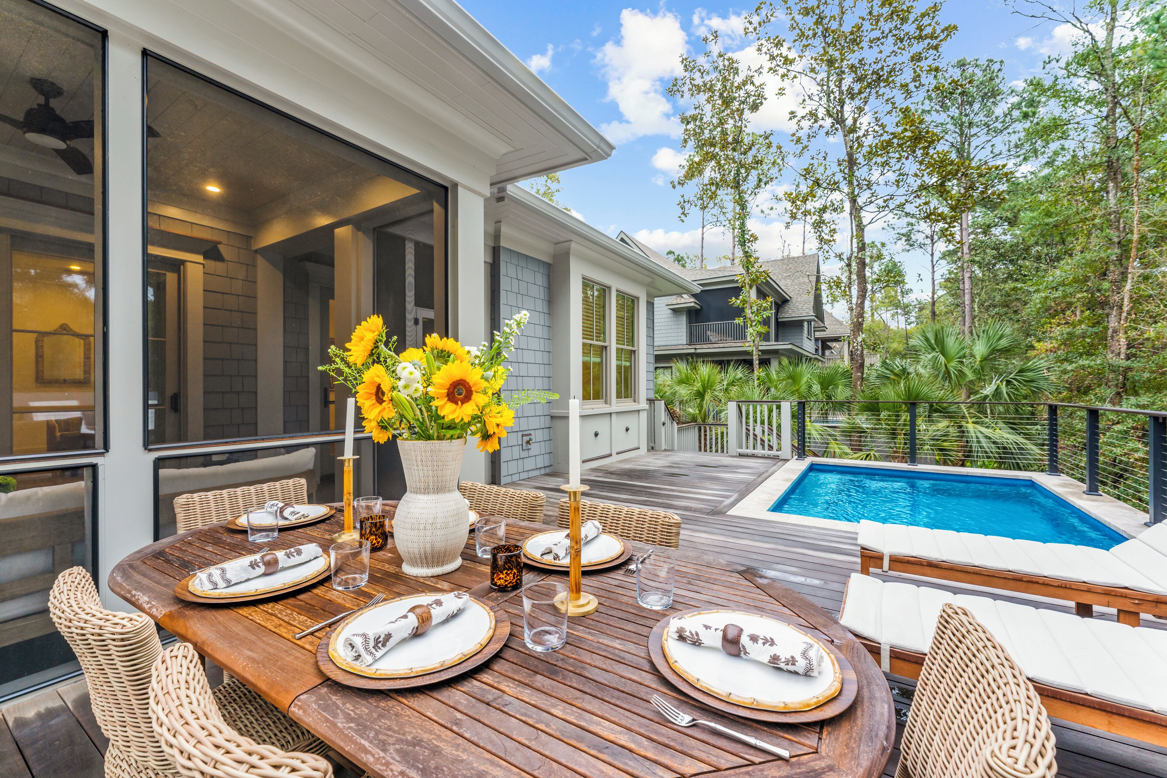 An outdoor dining area with a wooden table set for four, surrounded by wicker chairs, near a swimming pool.