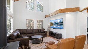 A spacious living room with a large sectional sofa, a round coffee table, and a leather armchair. There is a flat-screen TV mounted on the wall, and the room has large windows with blinds. - Shaver Escape – Hot Tub Cabin Retreat with AC + Forest Views (Shaver Lake)