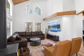 Ein geräumiges Wohnzimmer mit einer großen Eckcouch, einem runden Couchtisch und einem Ledersessel. Ein Flachbildfernseher ist an der Wand angebracht, und das Zimmer verfügt über große Fenster mit Jalousien.