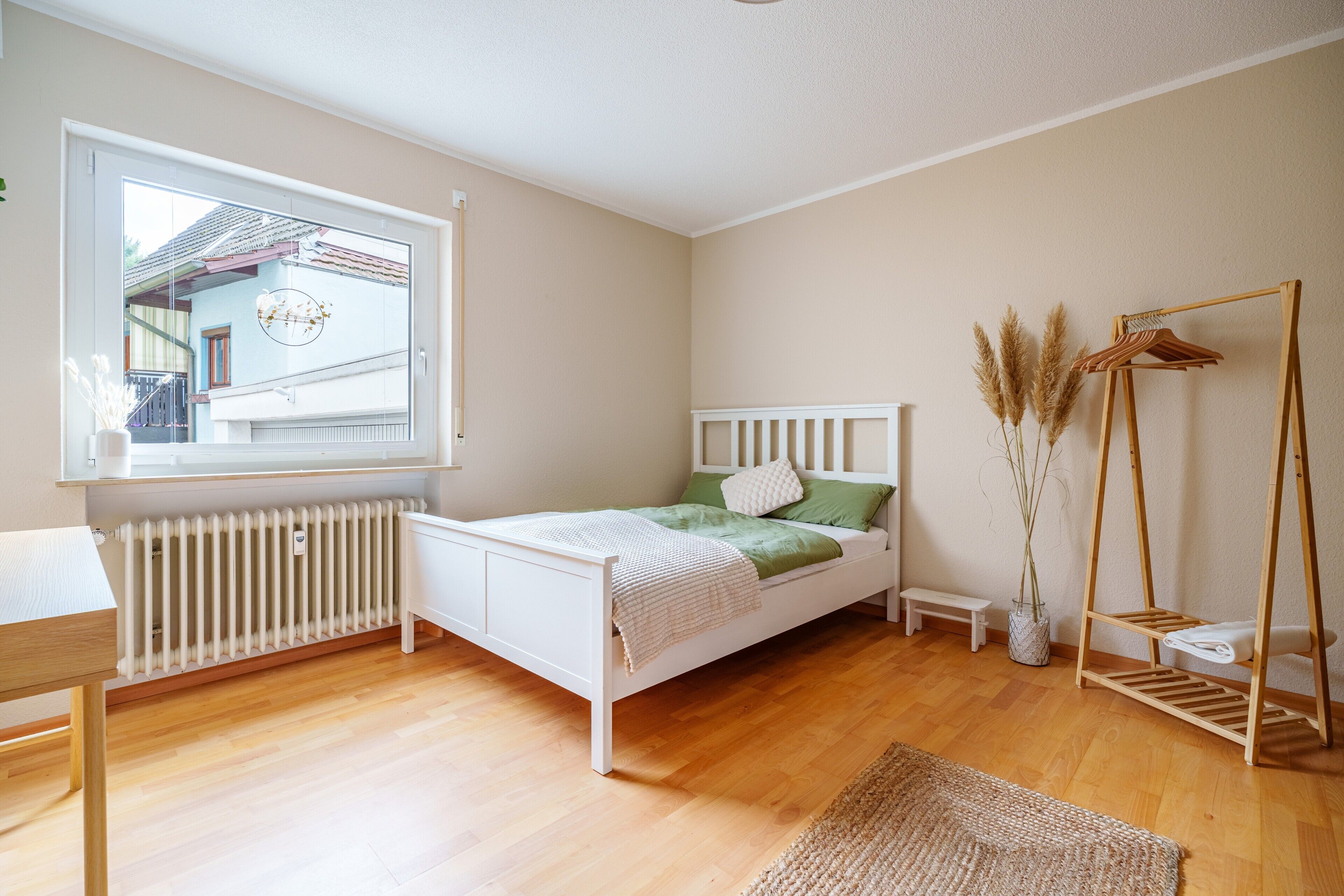 A bedroom with a white bed, a wooden floor, a window with a view, a bedside table, and a radiator.
