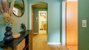 Un pasillo con un espejo, plantas decorativas, suelo de madera y una puerta que da a un baño.