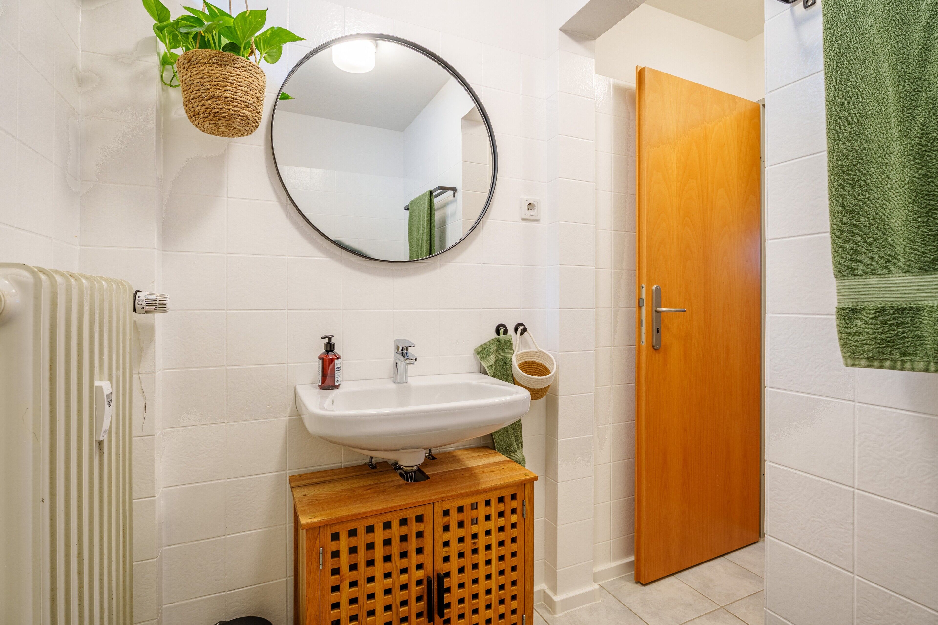 A bathroom with a white sink, a round mirror, a wooden cabinet, a green towel, and a wooden door.