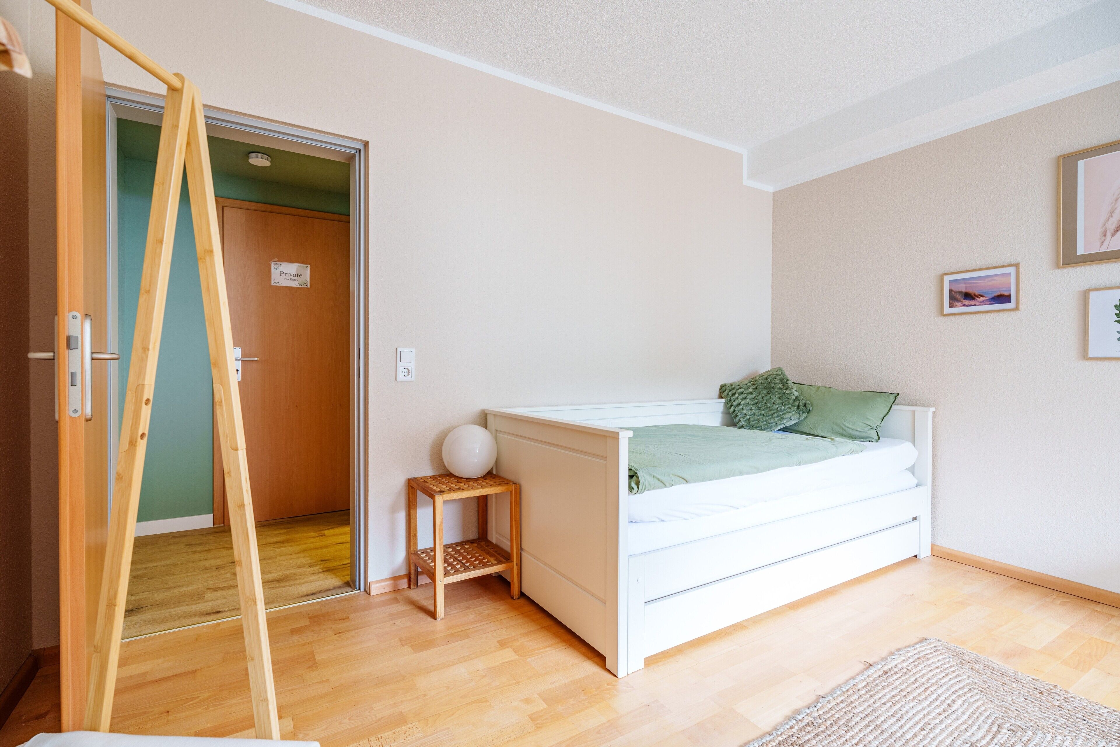 Un dormitorio con una cama blanca, un taburete de madera, un almohadón verde y un cuadro en la pared.
