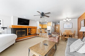 A modern living room with a fireplace, a ceiling fan, a dining area, and a kitchen in the background.