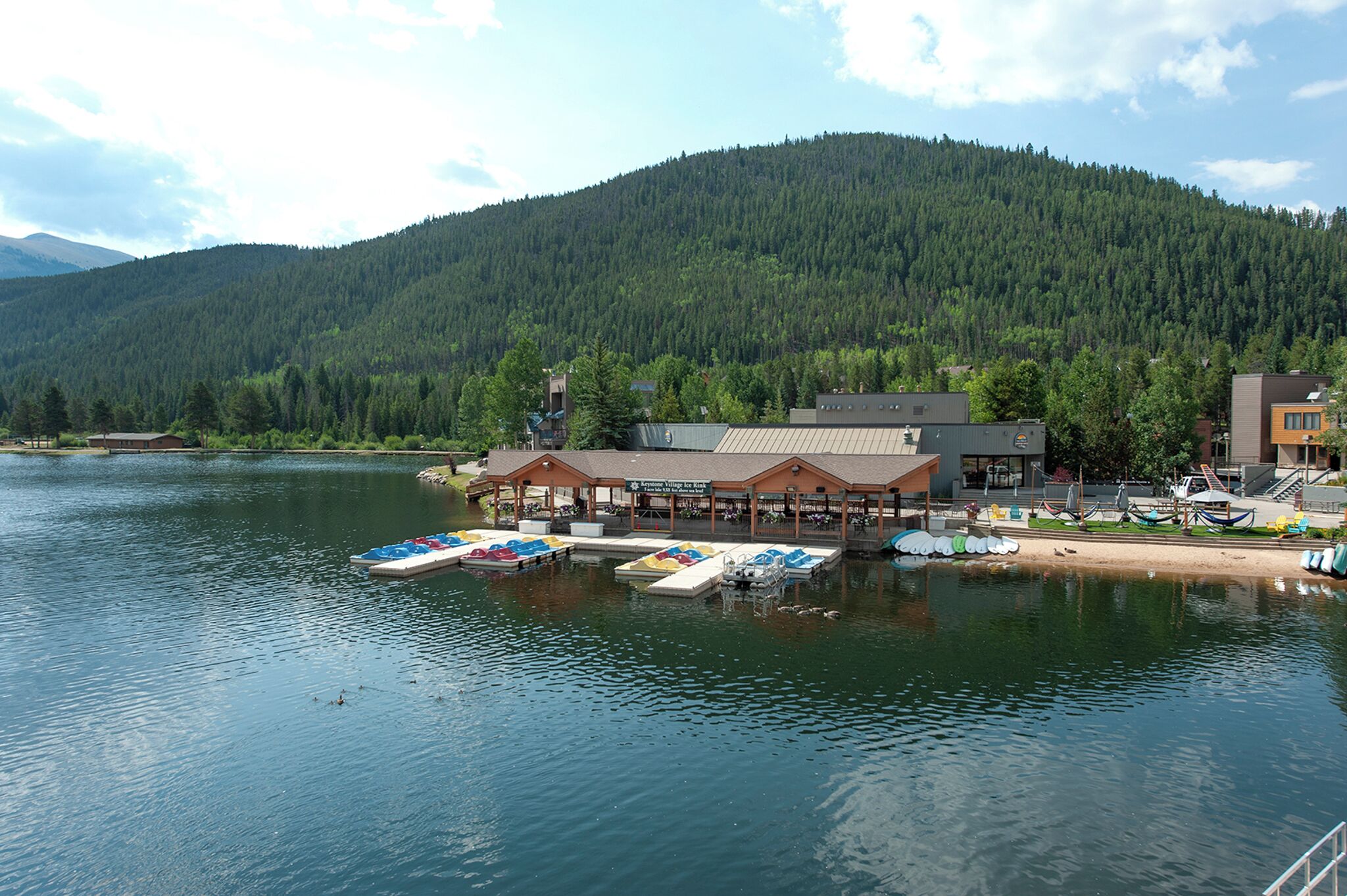 A lakeside building with a dock and boats, surrounded by forested hills.