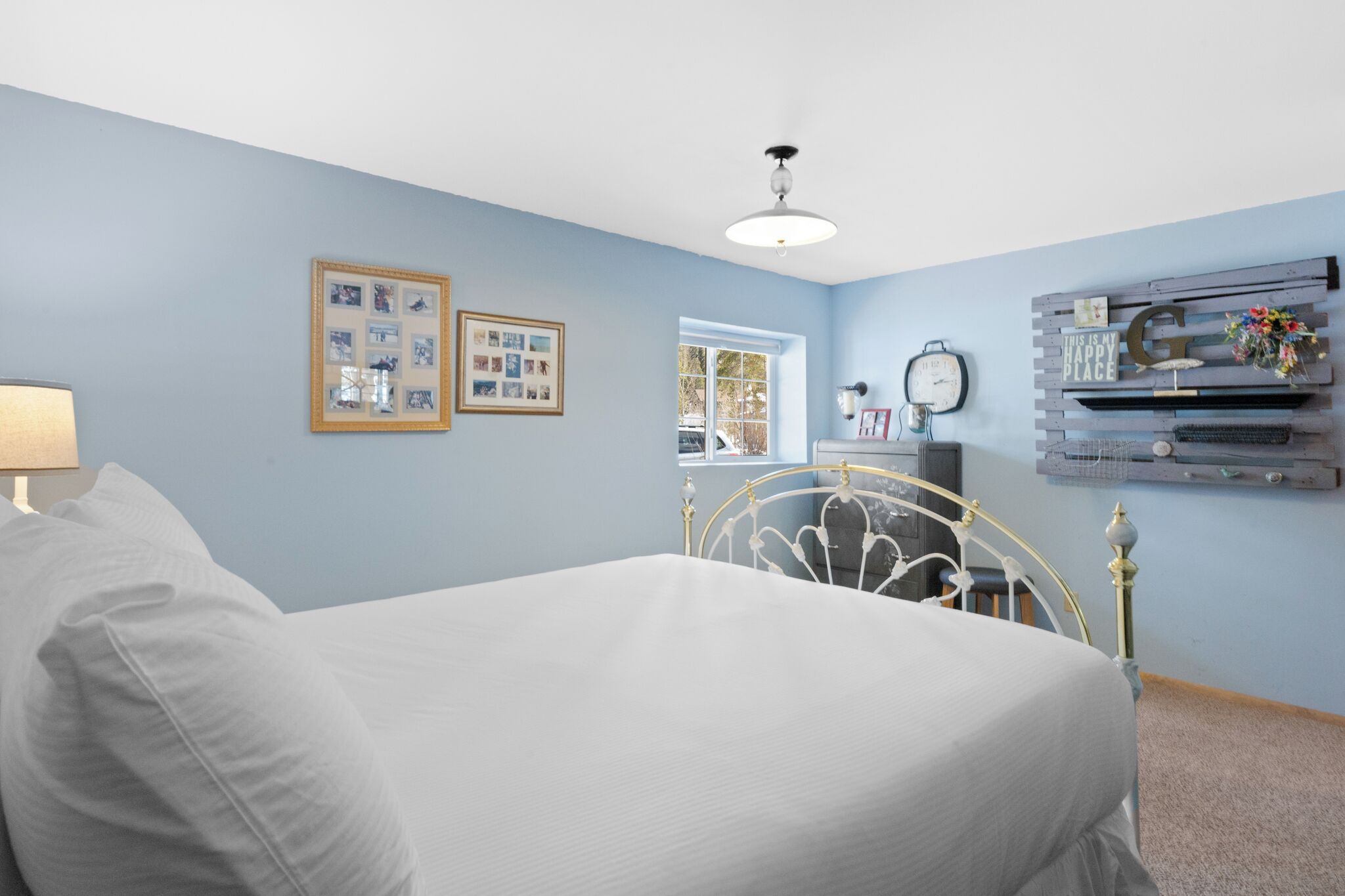 A bedroom with a bed, a window, a wall-mounted shelf with decorative items, and framed pictures on the wall.