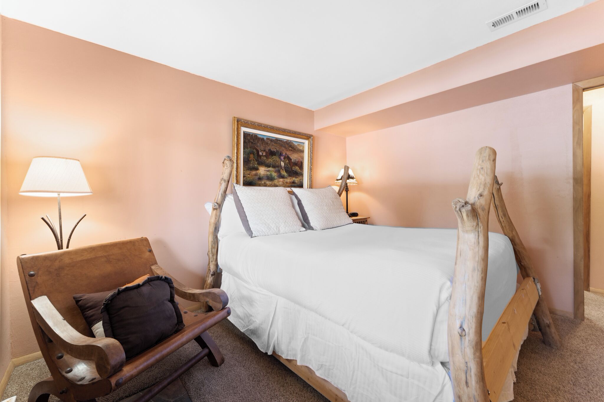 A bedroom with a wooden bed frame, a bench with cushions, a painting on the wall, and two bedside lamps.