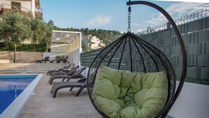 Una silla colgante junto a una piscina, con vista a un edificio y árboles al fondo.