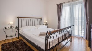 Un dormitorio con una cama, dos mesitas de noche, una araña y un ventanal con cortinas.