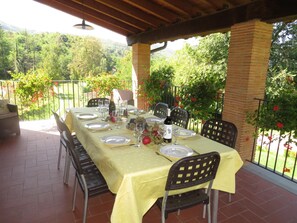 Un espace extérieur abrité, aménagé pour les repas, avec une table dressée pour quatre personnes, entourée de chaises, et offrant une vue sur un jardin luxuriant et des collines en arrière-plan.