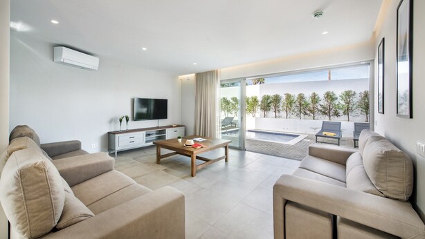 A modern living room with a flat-screen TV, sofas, a coffee table, and a sliding glass door leading to an outdoor area with a pool and greenery. - Luxury 6BR Villa Margarida | Pool, Hot Tub & Walk to Albufeira Strip (Albufeira)