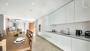 A modern kitchen with white cabinets, a wooden dining table, and a kitchen island. - Luxury 6BR Villa Margarida | Pool, Hot Tub & Walk to Albufeira Strip (Albufeira)