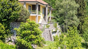 Ein zweigeschossiges Haus mit Balkon, umgeben von üppiger Vegetation und einer Steinmauer.