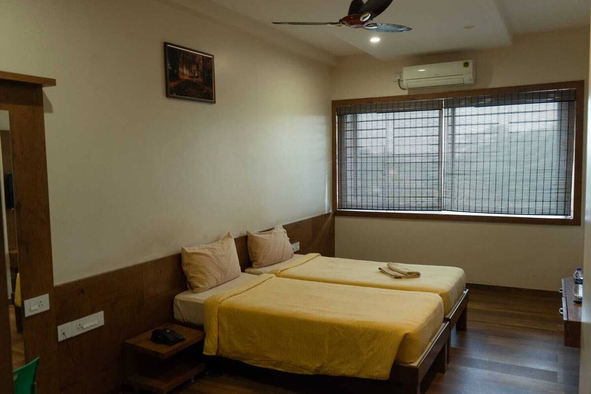 A room with two beds, a ceiling fan, a window with blinds, and a small table with a lamp.
