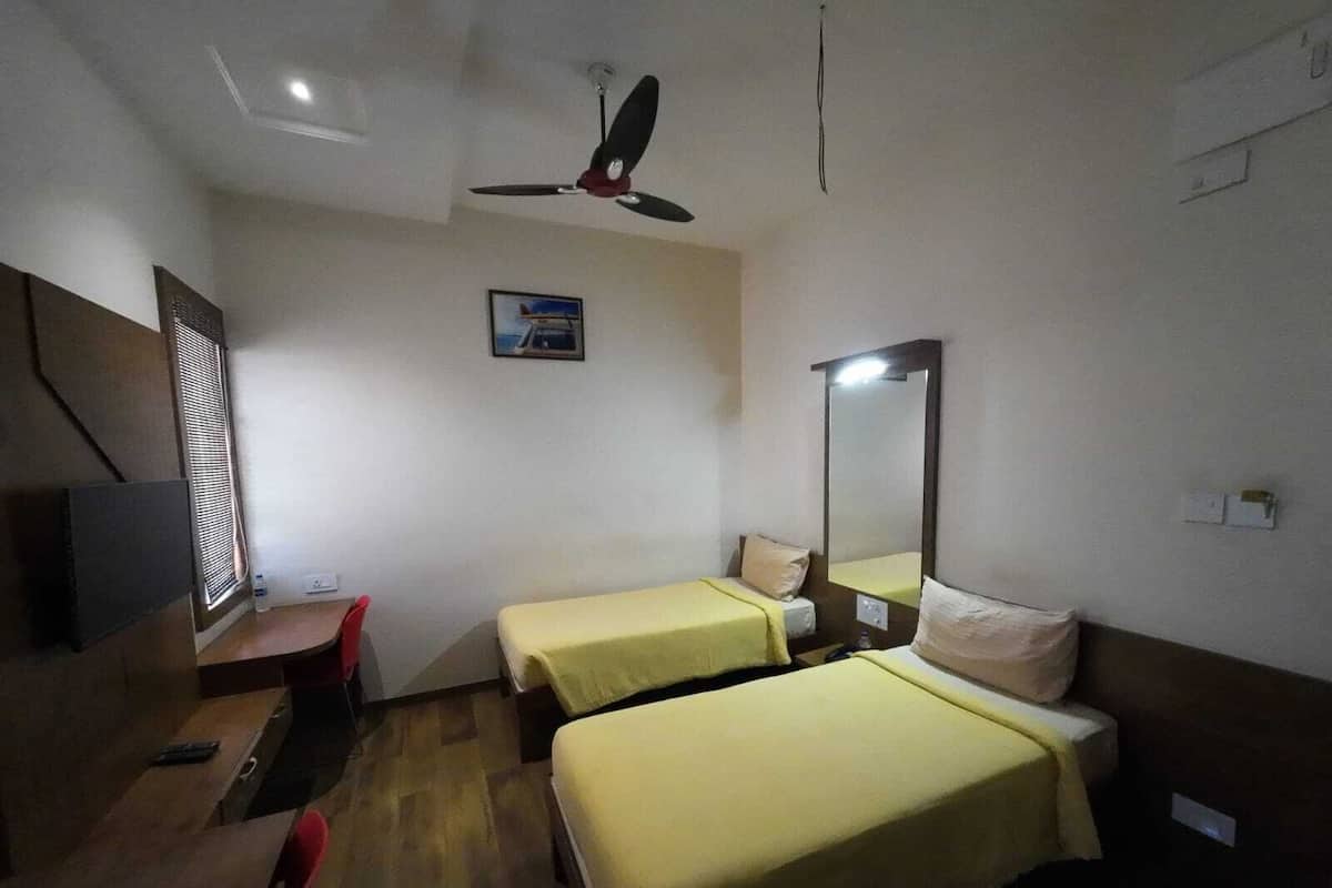 A hotel room with two single beds, a desk with a chair, a ceiling fan, a wall-mounted TV, and a window with blinds.