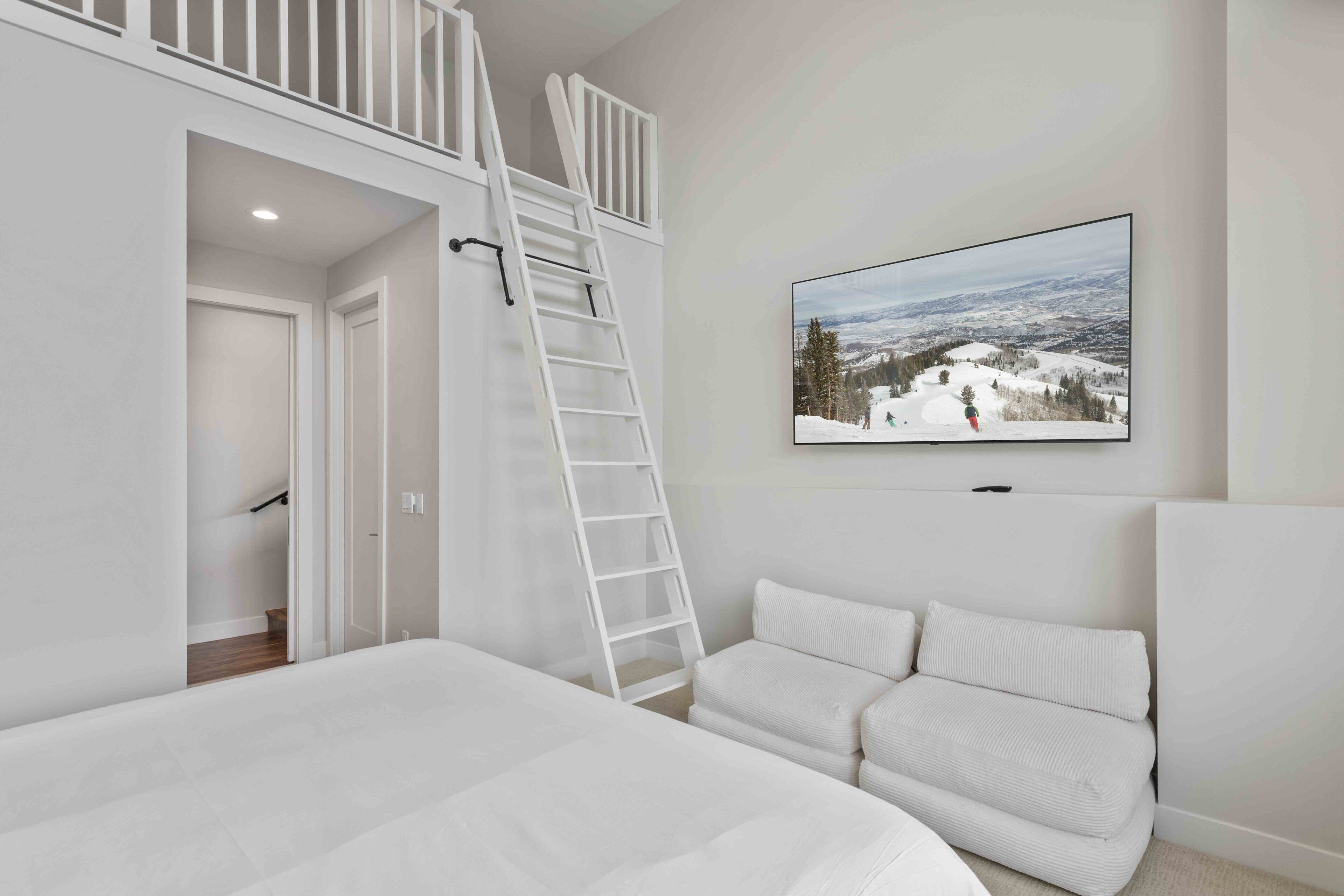 Une chambre avec un lit en mezzanine, une échelle blanche, un canapé blanc et un tableau représentant un paysage enneigé.