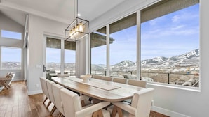 Un comedor moderno con una gran mesa de madera y sillas tapizadas en tono beige, con ventanas amplias que ofrecen vistas a montañas nevadas.