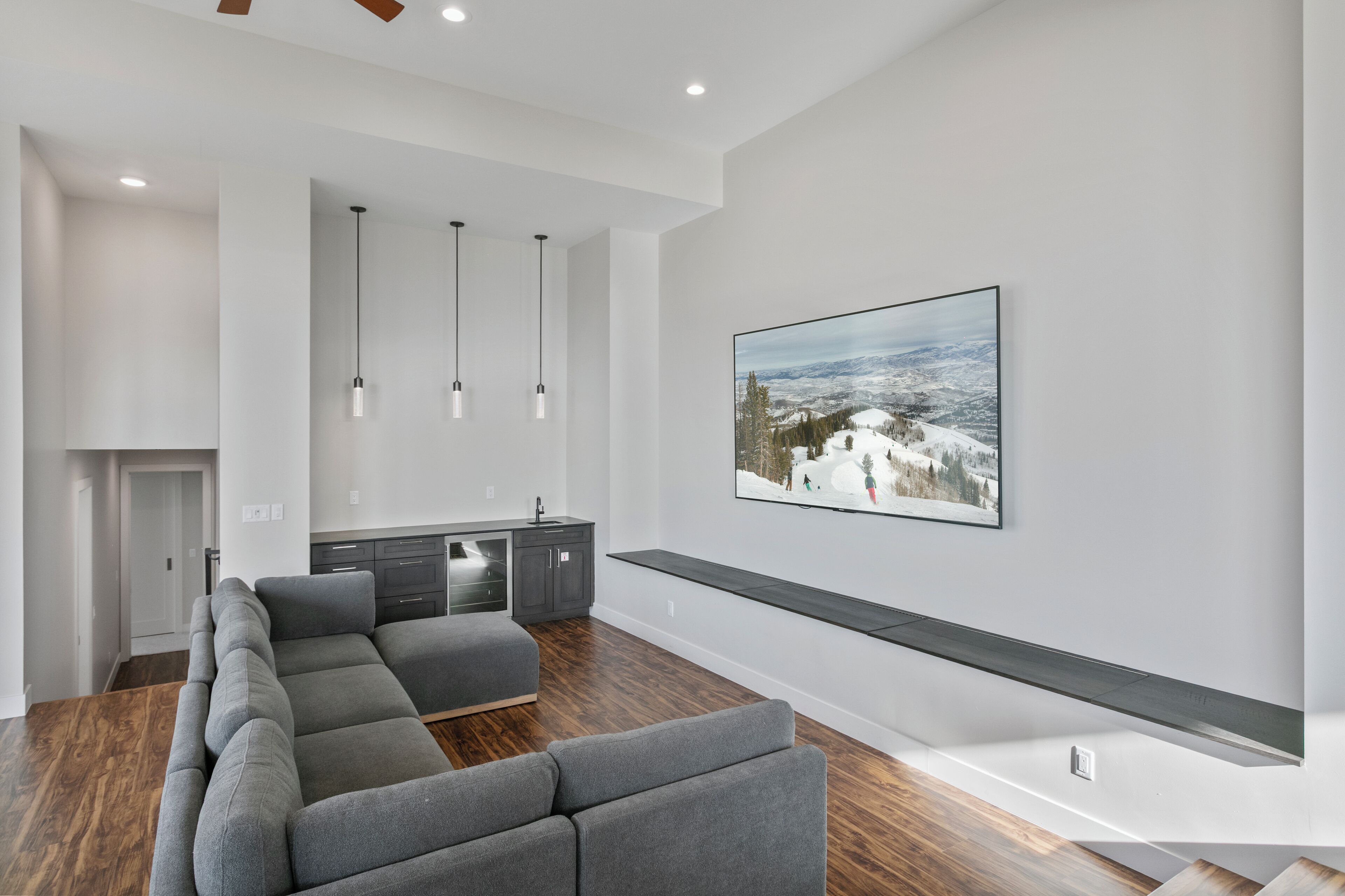 Una sala de estar moderna con un sofá modular, un televisor montado en la pared, lámparas colgantes y una cocina al fondo.