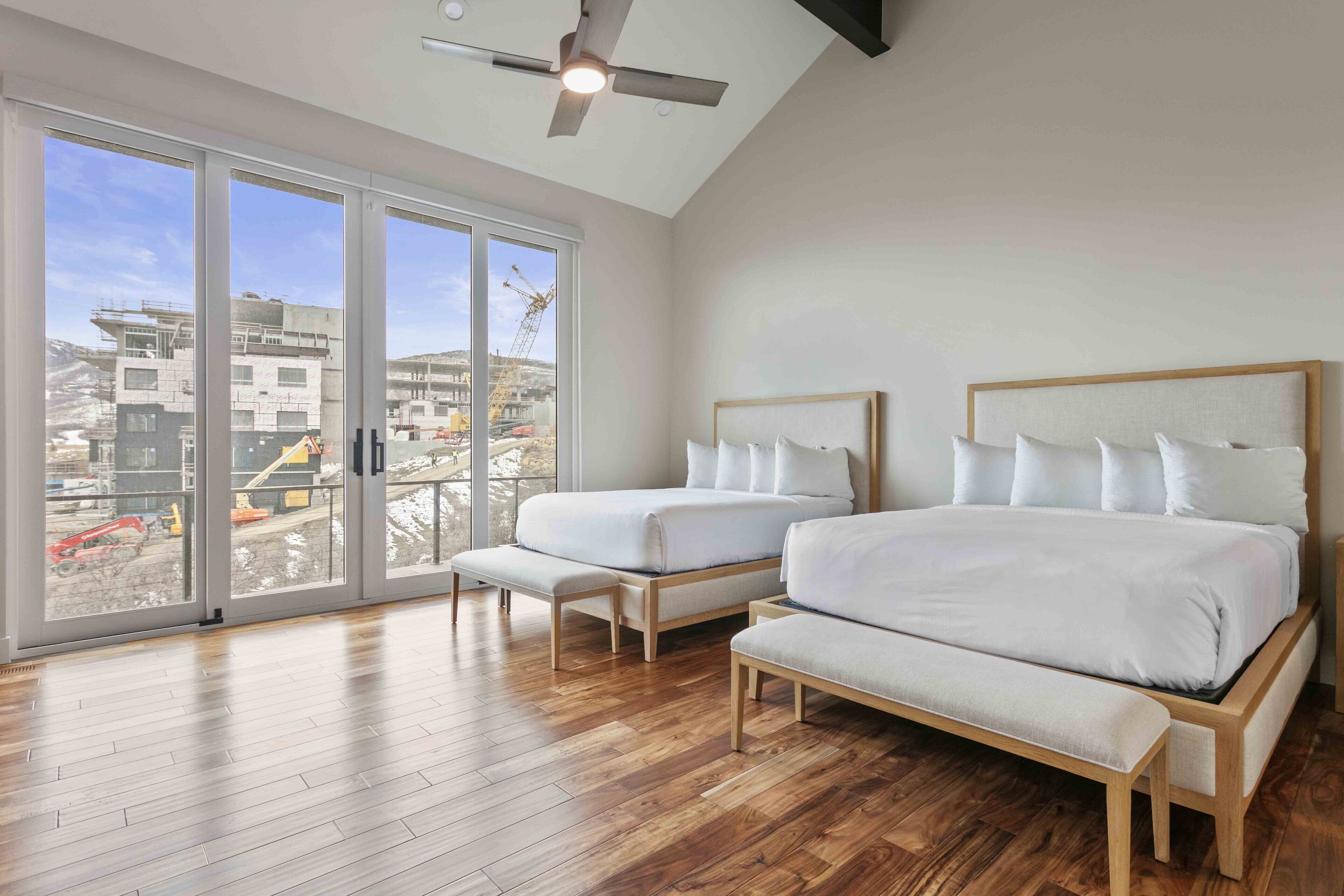 Une chambre avec deux lits, un ventilateur de plafond et de grandes fenêtres donnant sur un chantier de construction.