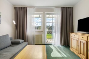 A room with a sofa, a wooden table, a TV, and a view through a door to an outdoor area. - Deluxe Apartment with Lake View K3 203 (Spalt)