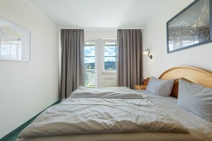 A hotel room with a bed, curtains, a nightstand, a wall-mounted lamp, and a window with a view. - Deluxe Apartment with Lake View K3 203 (Spalt)