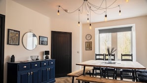 Un comedor con mesa de madera, bancos y sillas. Se observa un mueble azul oscuro con cajones y un espejo redondo en la pared. También hay un reloj y cuadros enmarcados.