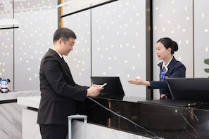 A reception desk with a man and a woman interacting, a panda plush toy on the counter, and a modern interior with a patterned wall. - Hampton by Hilton Ningde Lithium Electricity Town (Ningde)