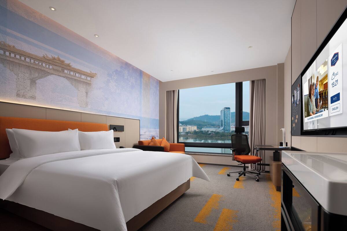 A hotel room with a large bed, a desk, a chair, a TV, and a view of a cityscape.