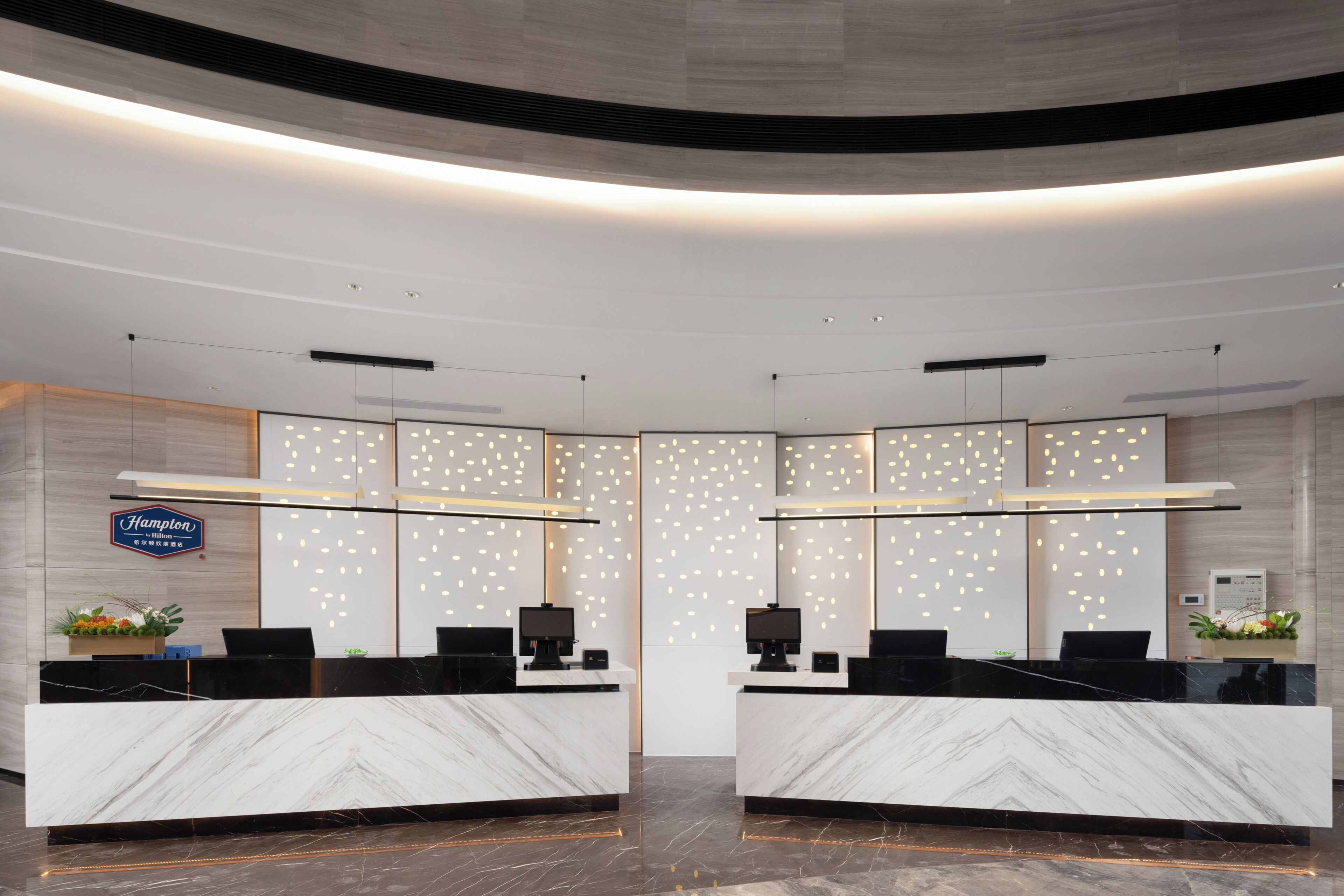 A hotel reception area with marble counters, modern lighting, and a sign indicating it's a Hampton by Hilton location.