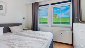 A bedroom with a bed, a dresser, a radiator, and a window with a view of a green field. - Dog-Friendly Apartment H1 005 (Spalt)