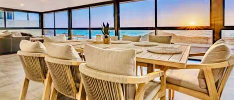 Amplia zona de estar al aire libre con sillas de mimbre y una mesa de madera, con vistas a una playa al atardecer.