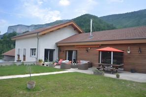 Una casa moderna con patio, mobiliario exterior y montañas al fondo.