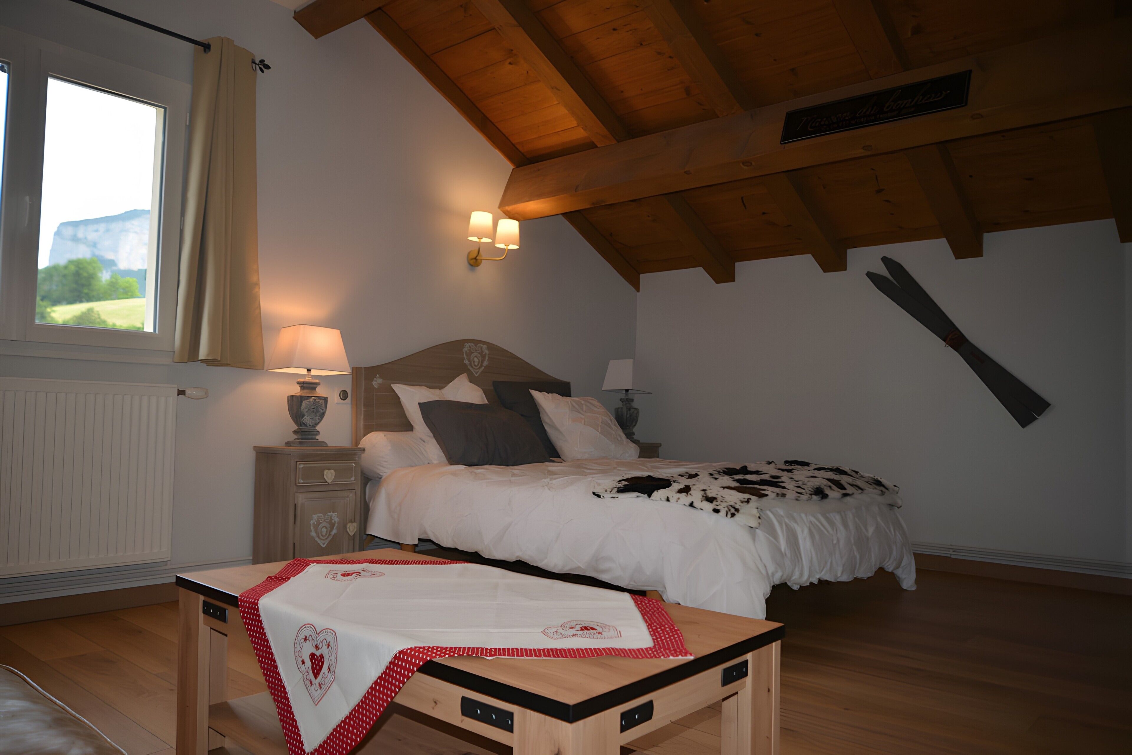 A bedroom with a wooden ceiling, a bed with a cowhide blanket, a bedside table with a lamp, and a window with curtains.