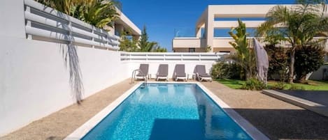 Una piscina rectangular con borde blanco, rodeada por un muro blanco y una zona de grava. Hay sillas de descanso alineadas a lo largo del muro.