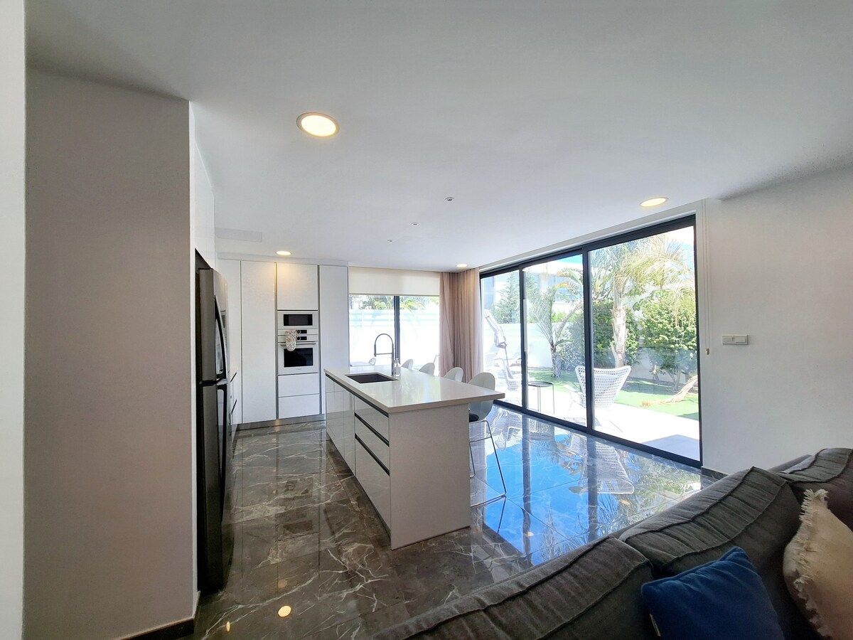 A modern kitchen with a central island, a dining area with a glass table, and a large sliding glass door leading to an outdoor space.